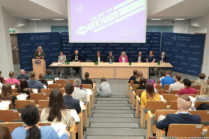 Dyktando Uniwersyteckie 2025. Sala wykładowa. Uczestnicy na siedzeniach tyłem do kadru. Za stołem 8-osobowe jury. Za mównicą po lewej prorektorka otwierająca wydarzenie. U góry ekran z logiem dyktanda w kształcie pióra do pisania, datą i godziną konkursu.