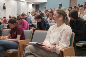 Dyktando Uniwersyteckie 2025. Sala wykładowa. Ujęcie bokiem na uczestników konkursu piszących dyktando.