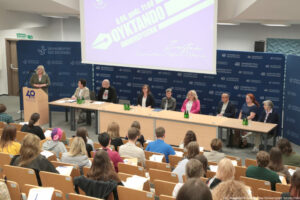 Dyktando Uniwersyteckie 2025. Sala wykładowa. Ujęcie bokiem. Uczestnicy na siedzeniach tyłem do kadru. Za stołem 8-osobowe jury. Za mównicą po lewej prorektorka otwierająca wydarzenie. U góry ekran z logiem dyktanda w kształcie pióra do pisania, datą i godziną konkursu.