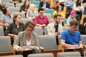 Dyktando Uniwersyteckie 2025. Sala wykładowa. Ujęcie na uczestników konkursu piszących dyktando. Na pierwszym planie dziewczyna w różowo-fioletowych włosach.