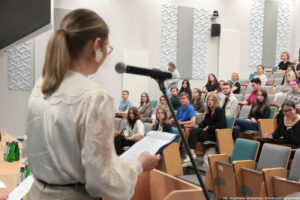 Dyktando Uniwersyteckie 2025. Sala wykładowa. Ujęcie bokiem. Na pierwszym planie z lewej strony tyłem do kadru prowadząca, stojąca za mównicą z mikrofonem na statywie. W tle uczestnicy na siedzeniach.