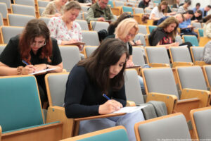 Dyktando Uniwersyteckie 2025. Sala wykładowa. Ujęcie bokiem na uczestników konkursu piszących dyktando.